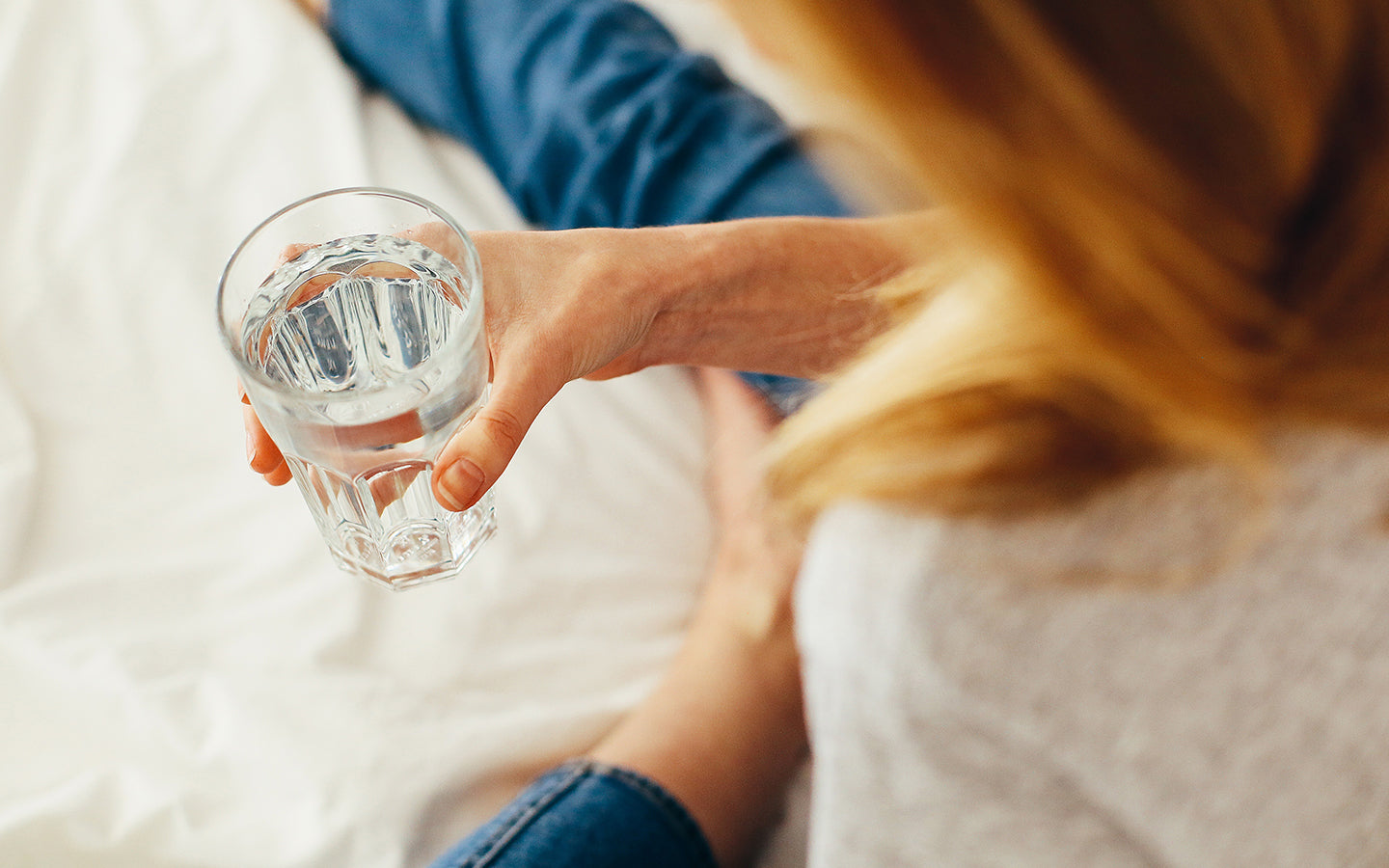 woman drinking a glass of water while sitting on bedsheets