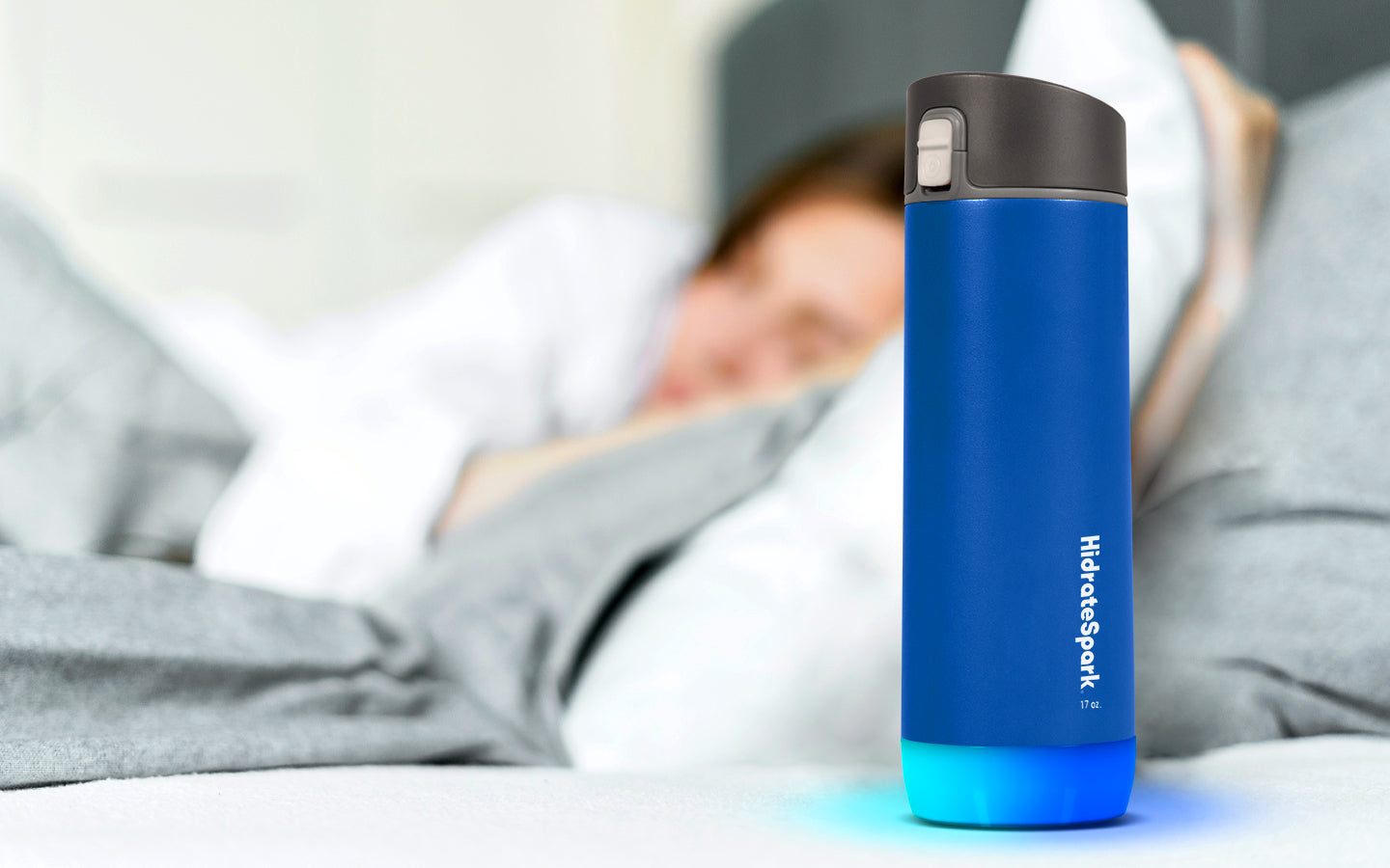 A Deep Blue 17 oz HidrateSpark smart water bottle sits in front of a woman lying in bed meant to prevent dehydration during sleep..