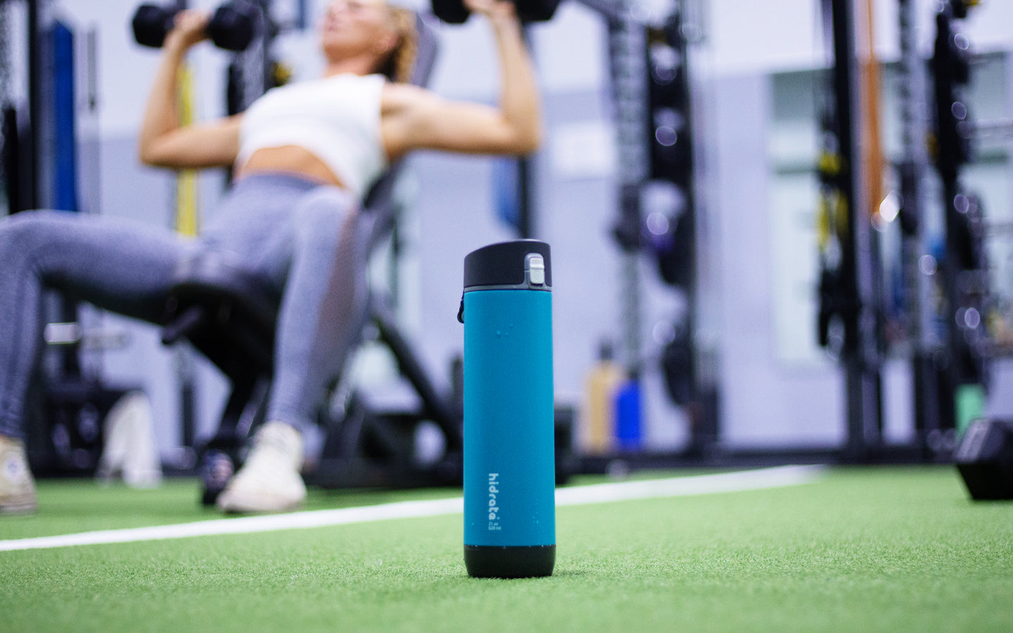 A 21 oz Seaglass color HidrateSpark smart water bottle sits in the foreground while a woman exercises in the background.